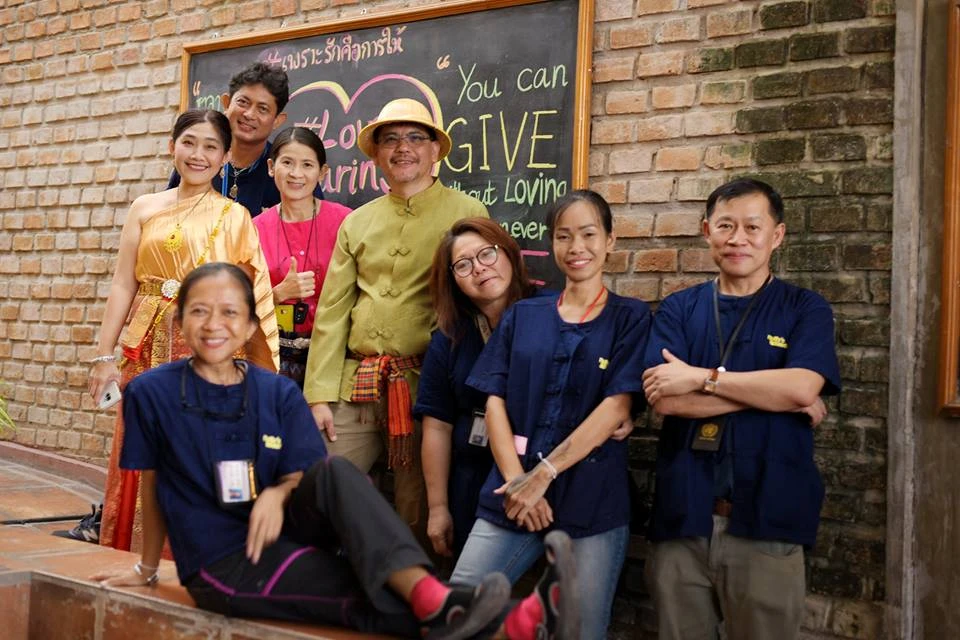 A diverse team of eight people, including staff in traditional Thai attire and navy shirts, posing in front of a chalkboard with a message about giving