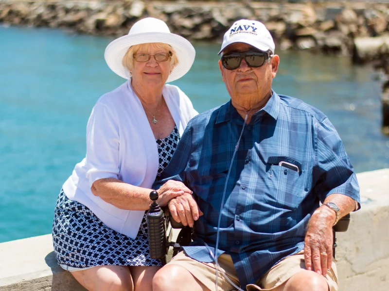 Elderly couple sitting by the water, holding hands, with one person using a mobility scooter and oxygen support
