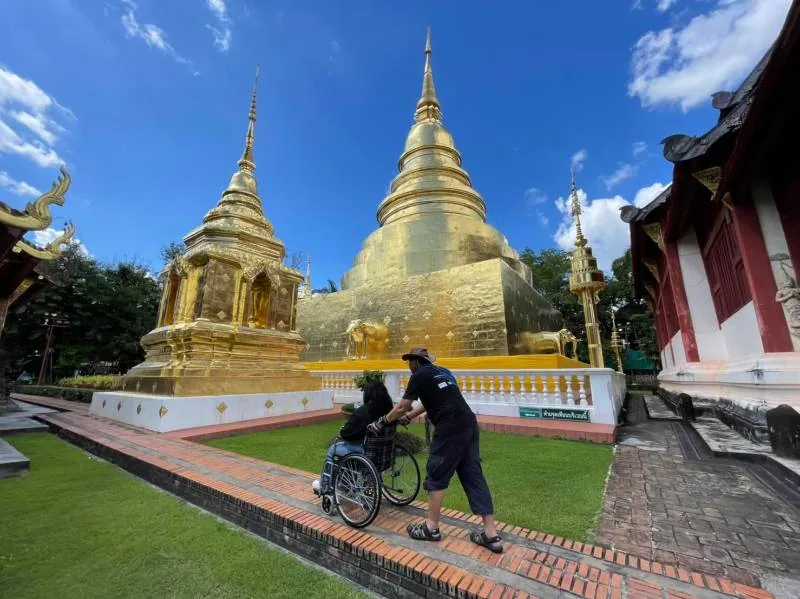A person in a wheelchair being assisted along a brick path near a golden stupa at a Thai temple