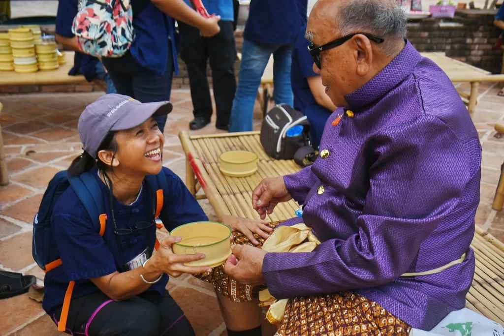 A local guide trainee kneeling and smiling while offering a food container to an elder in traditional attire