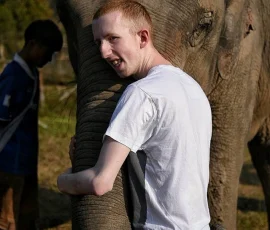 A memory that touches the soul! Experience the unique texture and warmth of an elephant's trunk through a close, gentle hug at our responsible sanctuary visit. A young male traveller, likely visually impaired, smiles warmly while embracing the thick, textured trunk of a friendly elephant at an ethical sanctuary in Chiang Mai, showcasing the tactile connection between human and animal.