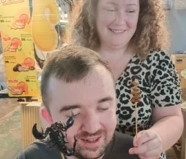 Exploring the unique textures and aromas of Chiang Mai’s street food during a sensory-focused night market adventure A blind male traveler wearing an "independence" t-shirt exploring a fried scorpion on a stick through touch and smell, accompanied by a smiling woman at a Chiang Mai night market.