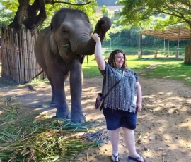 Pure joy and a gentle connection! Sharing a playful moment with one of the young elephants, an unforgettable highlight that engages all the senses. A joyful female traveller, with a wide smile, stands close to a baby elephant which playfully raises its trunk, showcasing a safe, tactile, and highly engaging interaction at a Chiang Mai elephant sanctuary.
