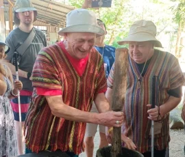 Cultural immersion meets accessible fun! Don traditional attire and enjoy the tactile experience of pounding ingredients to prepare a special treat for our gentle giants. A happy male visually impaired traveler, dressed in a traditional Thai pattern shirt, actively using a large wooden pestle to pound ingredients in a mortar, preparing food for the elephants at a Chiang Mai sanctuary, alongside another traveller with a white cane.