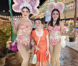 Experiencing the vibrant colors and friendly smiles of the Chiang Mai Night Bazaar. A blind female traveler smiling while standing between two Thai ladyboy performers in elaborate pink butterfly costumes at a Chiang Mai night market.