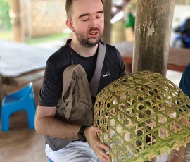The rewarding feeling of exploring Thai heritage through traditional handicrafts. A blind man smiling while holding and feeling the texture of a large, hand-woven bamboo basket in an outdoor workshop.