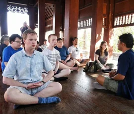 Finding inner peace through guided meditation in a traditional wooden pavilion. A group of young men and women sitting in a circle on a wooden floor in a traditional Thai pavilion, practicing meditation with a teacher.