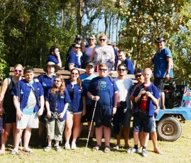 Exploring the charm of Baan Dok Bua village on our local farm vehicle tour. A group of international travelers, including those with visual impairments using white canes, posing together by a blue local farm vehicle in a sunny village setting.
