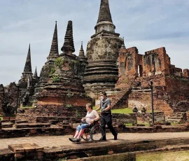 Inclusive heritage experiences in Thailand—where ancient beauty meets modern accessibility A wheelchair user being assisted along a paved path through ancient brick stupas at a heritage site
